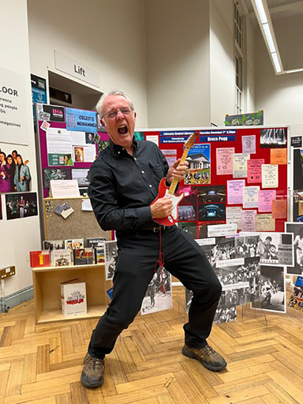 Bruce Pegg at the central library making a rock pose and playing an electrical guitar in front of a stand. 